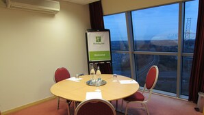 A conference room with a round table, four chairs, a projector screen displaying 'Holiday Inn Welcome', and a view of a bridge and buildings through large windows. - Holiday Inn Birmingham North - Cannock by IHG (Cannock)