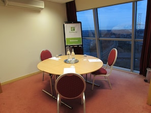 A conference room with a round table, four chairs, a projector screen displaying 'Holiday Inn Welcome', and a view of a bridge and buildings through large windows. - Holiday Inn Birmingham North - Cannock by IHG (Cannock)