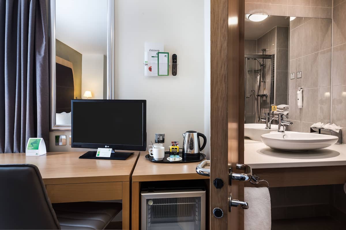 A hotel room with a desk, a flat-screen TV, a coffee maker, a kettle, a remote control, and a bathroom with a sink and shower.