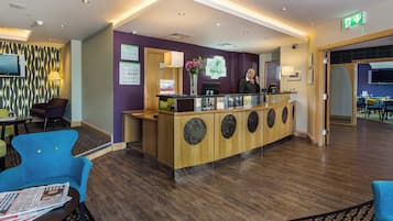 A hotel reception area with a wooden reception desk, a flat-screen TV, and a seating area with chairs.