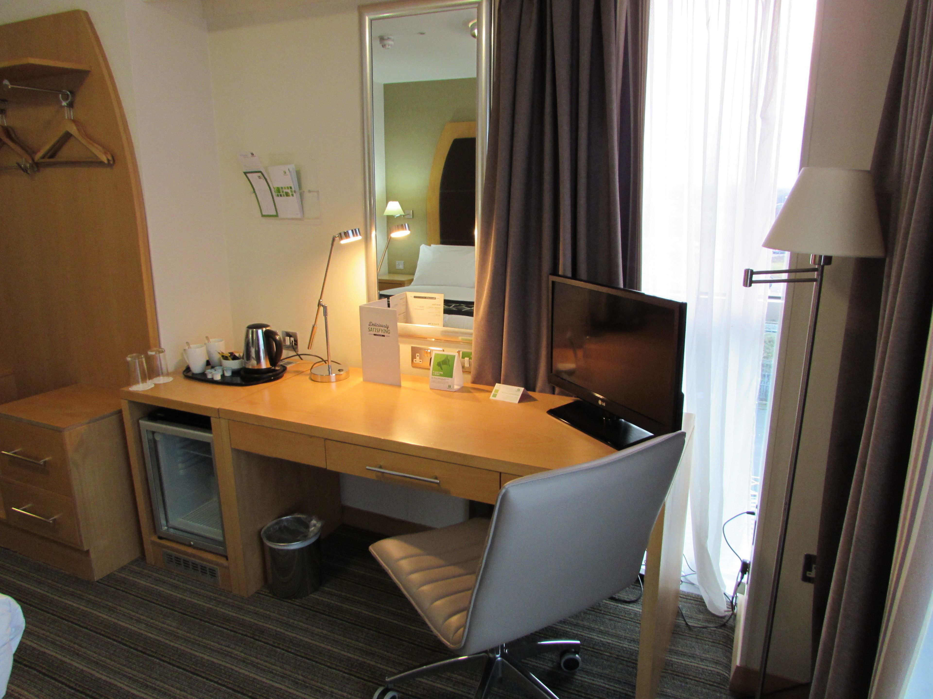Habitación de hotel con escritorio, una silla, televisor, lámpara, cubo de basura y espejo.