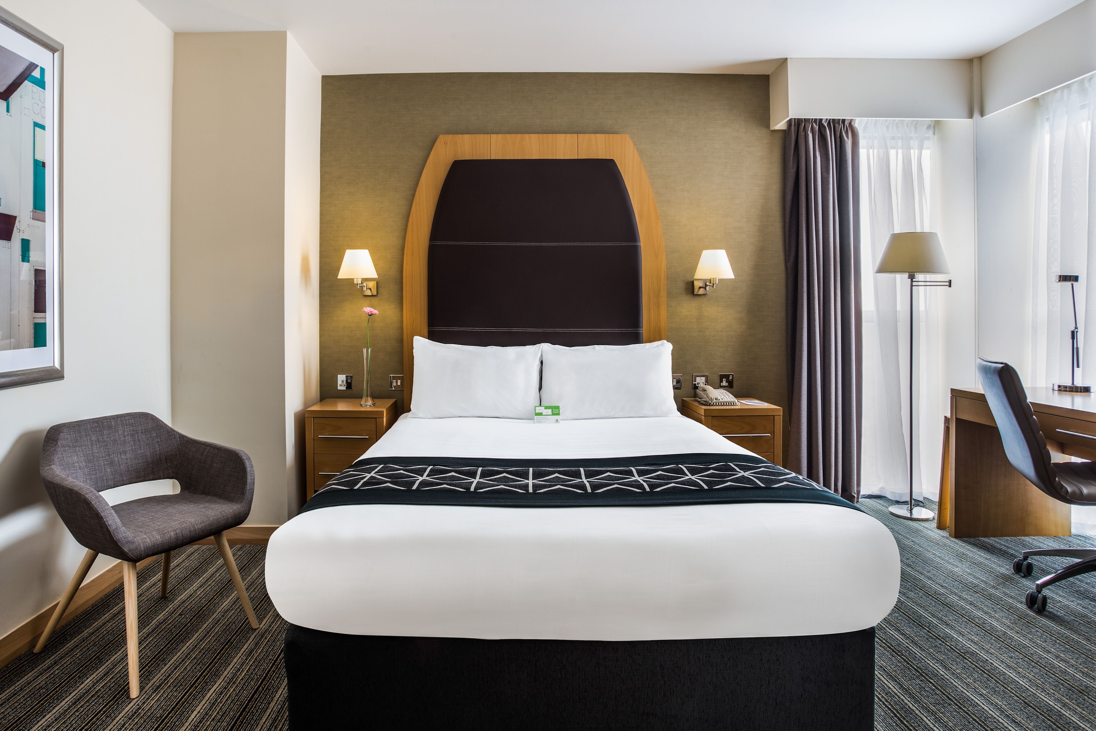 A hotel room with a large bed, two bedside tables with lamps, a desk, a chair, and a large window with curtains.