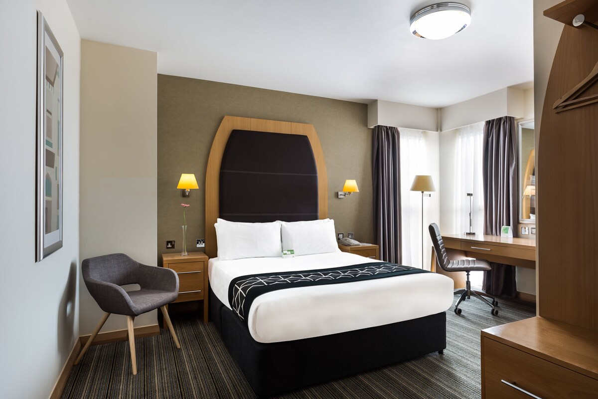 Une chambre d’hôtel avec un grand lit, un bureau, une chaise et une petite table avec une lampe.