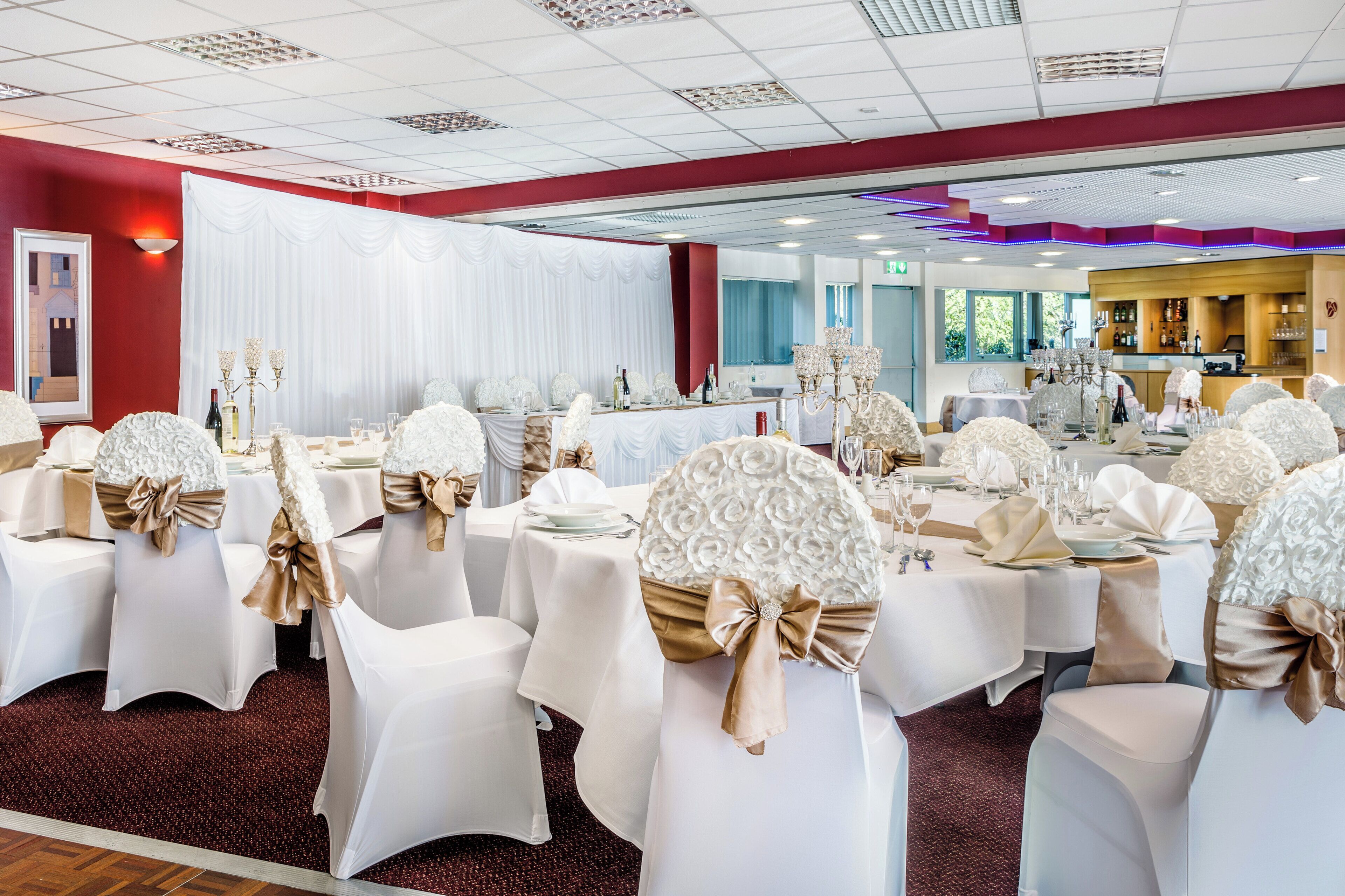 Une salle de banquet avec des tables rondes, des nappes blanches et des chaises recouvertes d’housse blanche et agrémentées de rubans dorés.