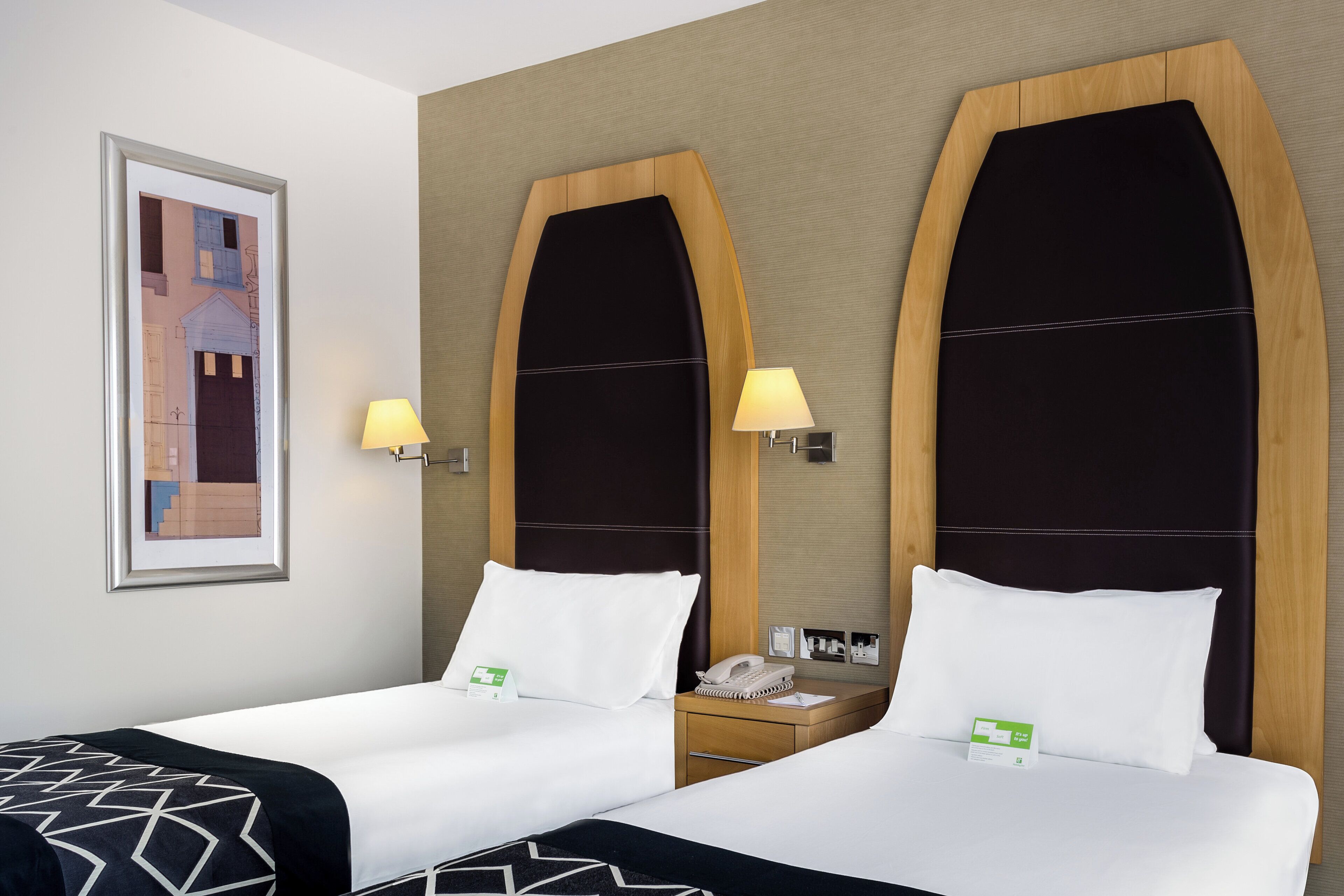 A hotel room with two beds, a framed picture on the wall, and a nightstand with a lamp and phone.