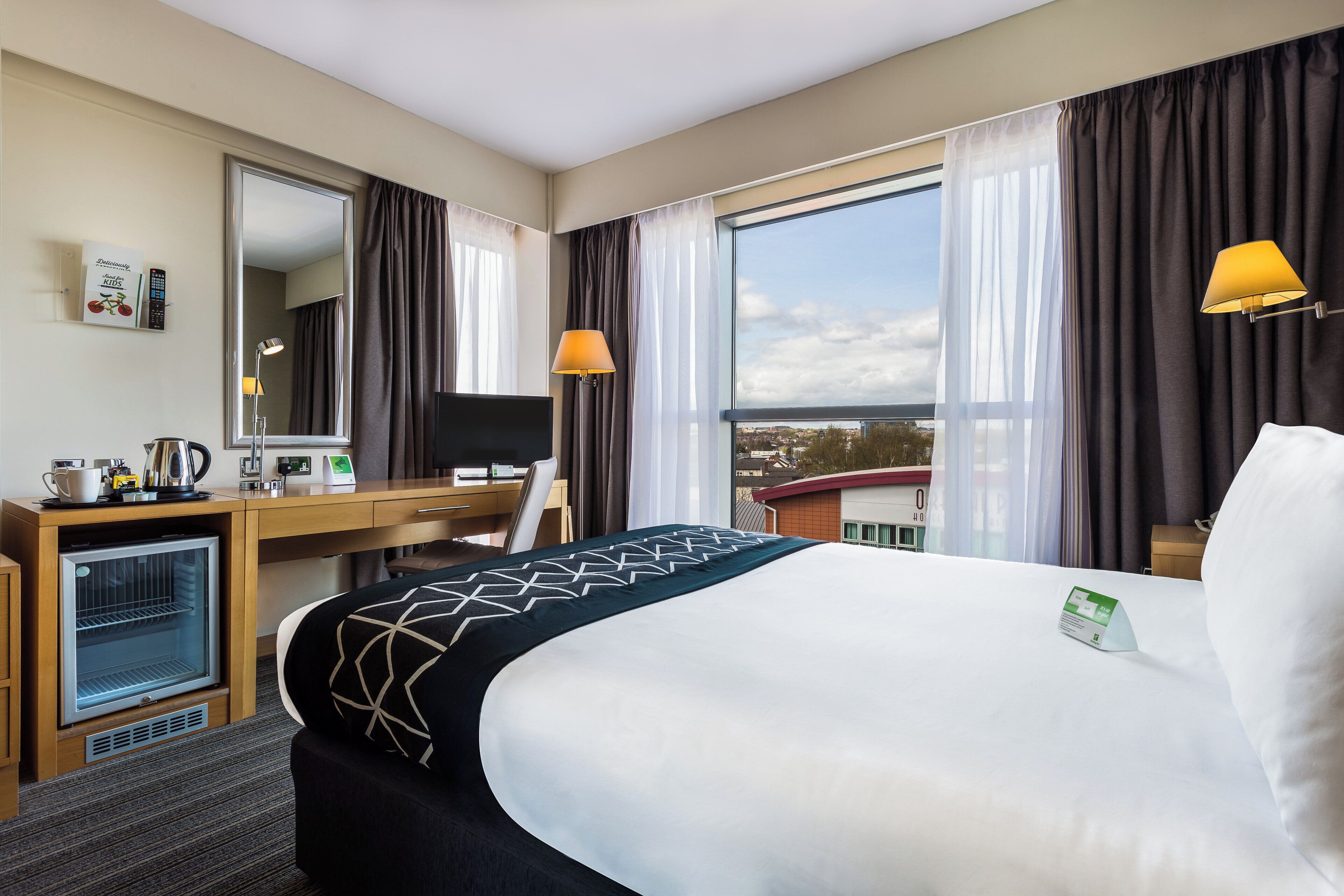 A hotel room with a bed, desk, refrigerator, and a large window with curtains.