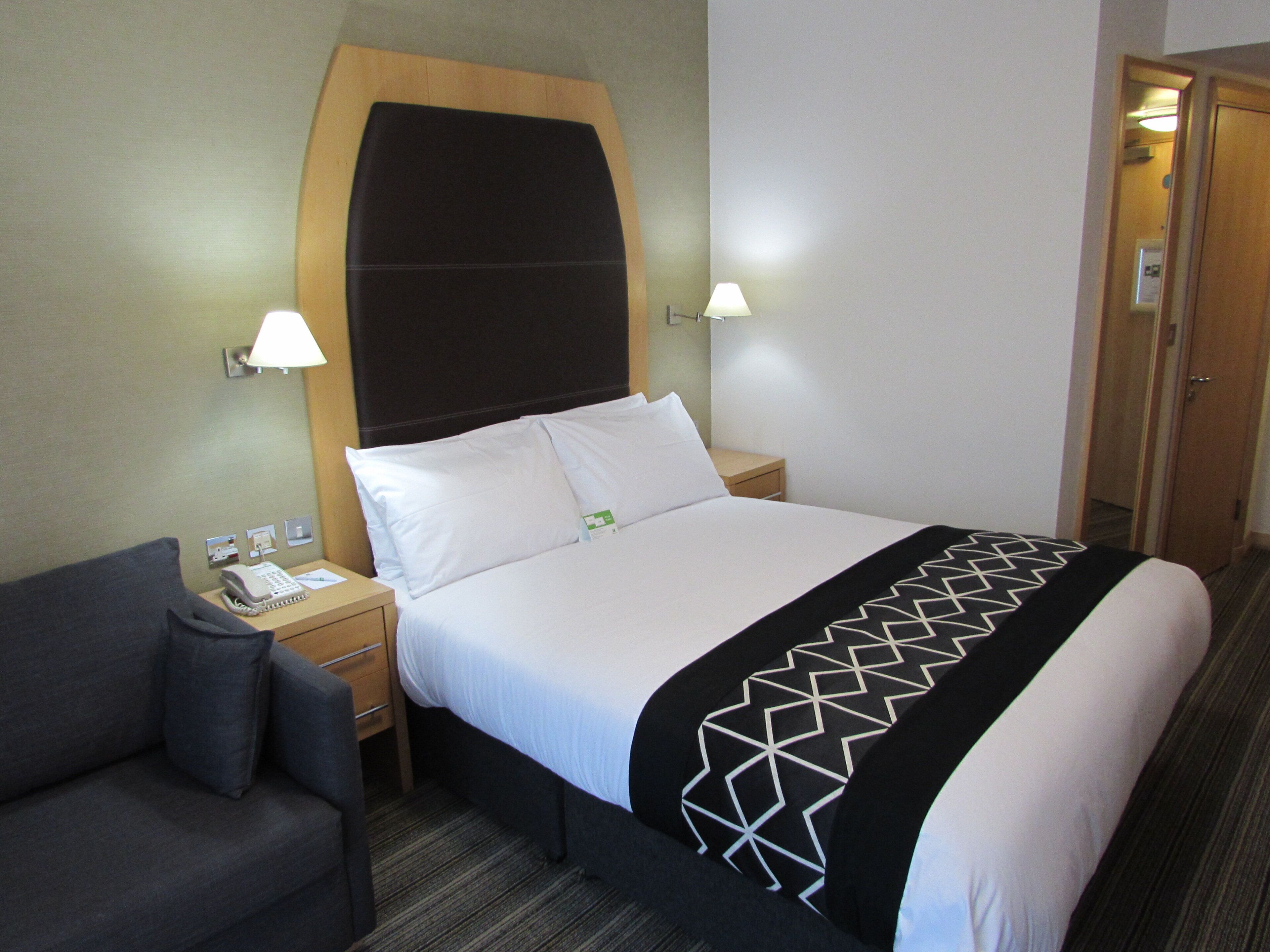A hotel room with a bed, bedside lamps, a telephone, a chair, and a door.