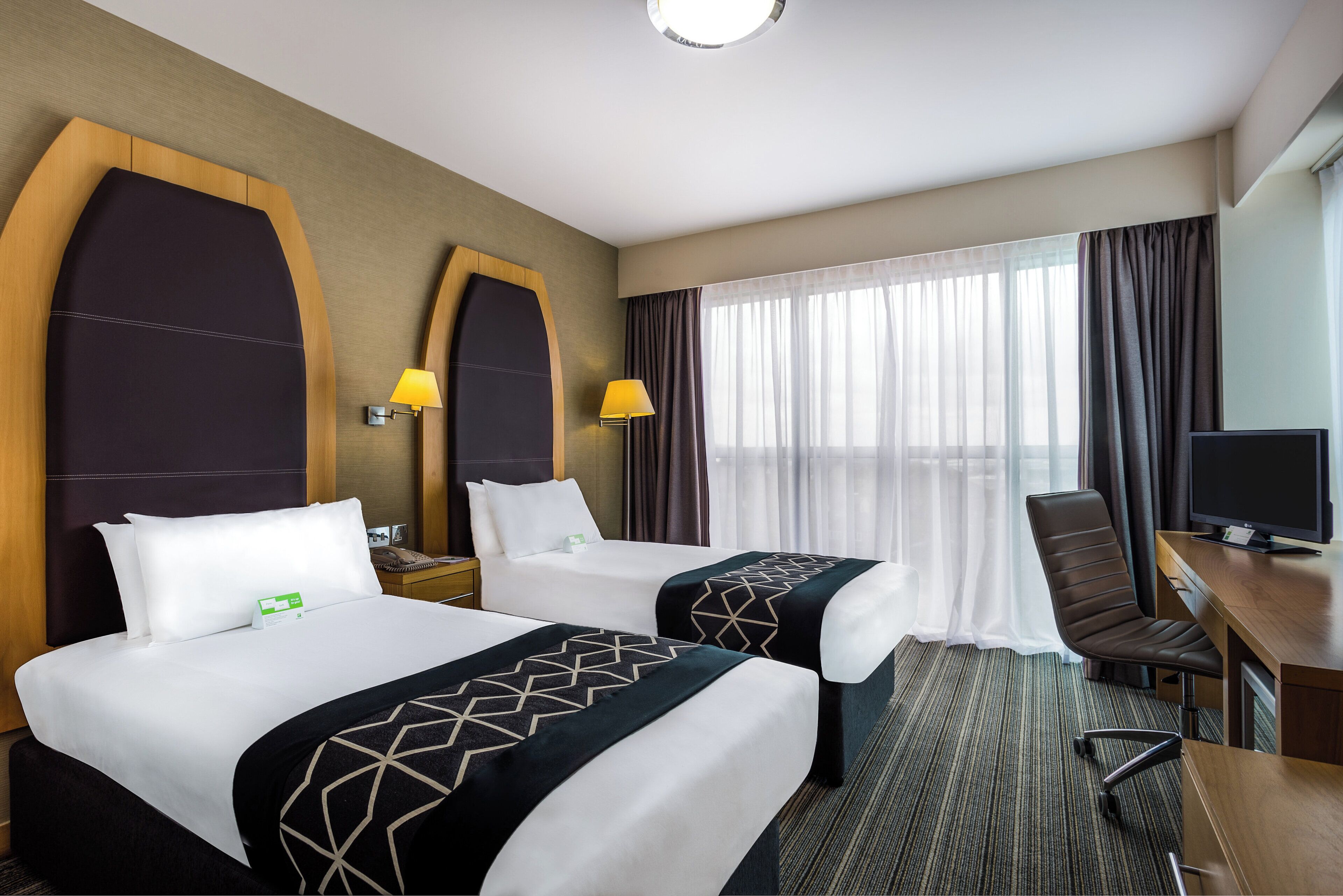 A hotel room with two beds, a desk, a chair, a TV, and a window with curtains.