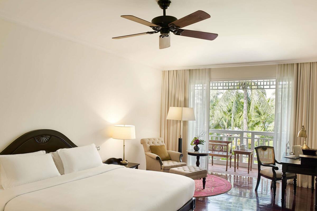 Habitación de hotel con cama, escritorio, silla, mesa y balcón con vista a palmeras.