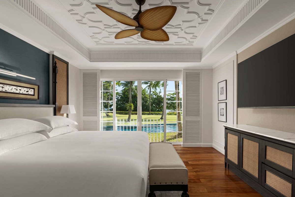 Ein Schlafzimmer mit Bett, einer Bank, einem Deckenventilator, einem Fernseher und Blick auf ein Schwimmbecken und Grünflächen.