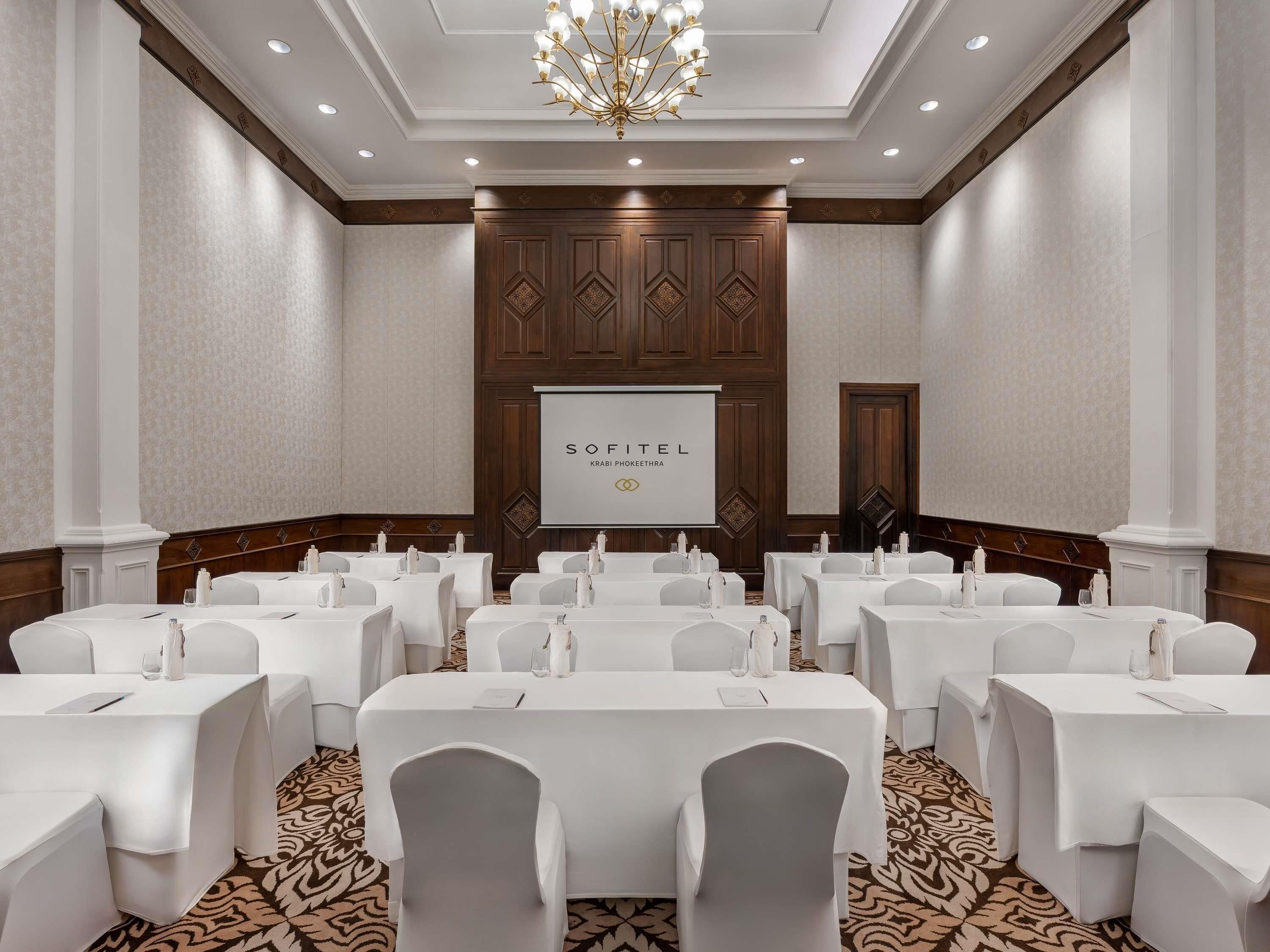 A conference room with multiple tables, white chairs, a projector screen, and a chandelier.