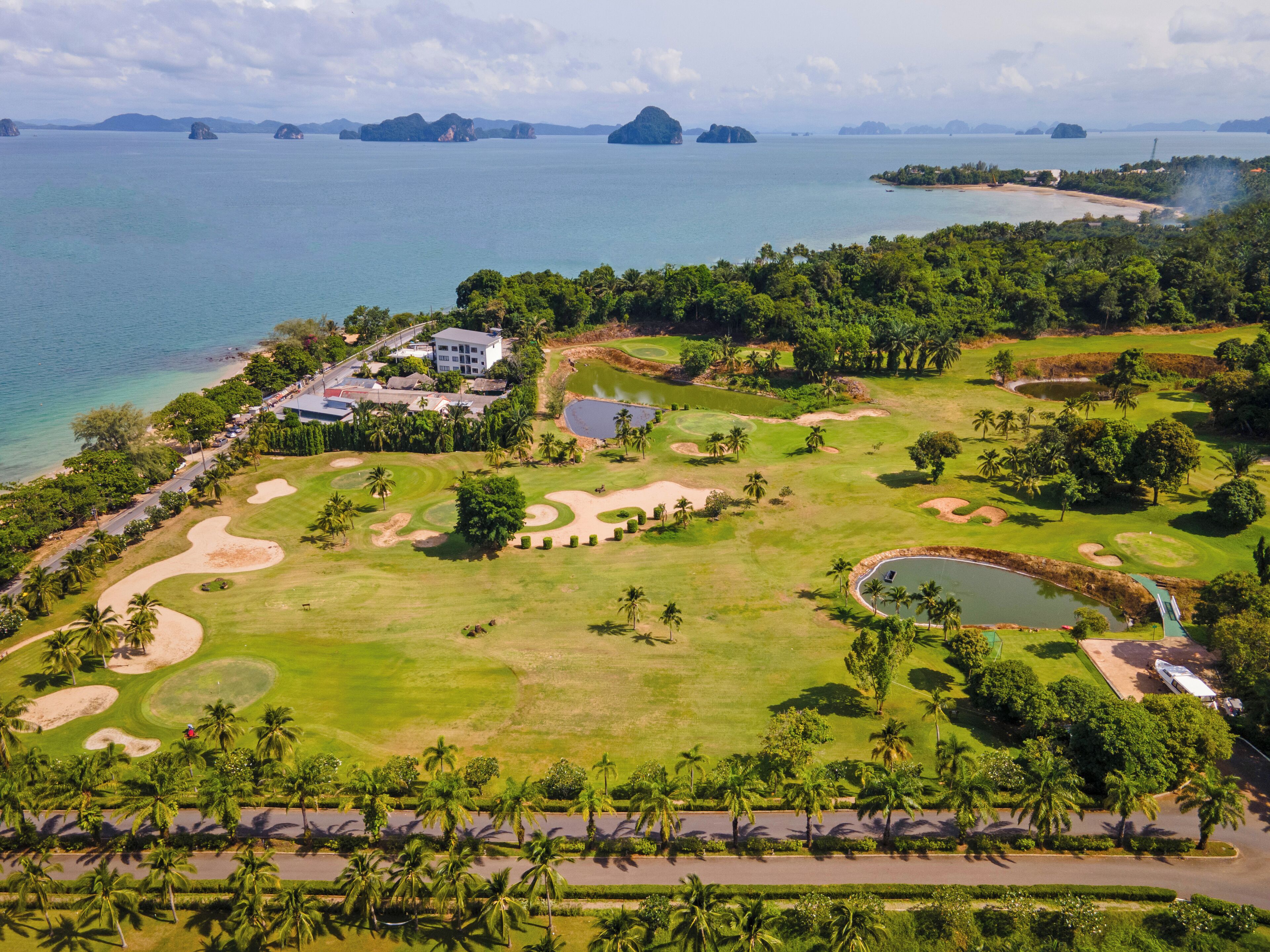 Vista aérea de un campo de golf con un club de campo, obstáculos acuáticos y palmeras.