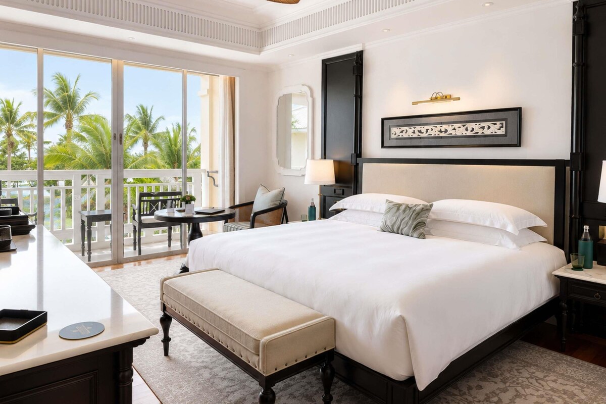 Une chambre d’hôtel avec un grand lit, un bureau avec une chaise, un balcon avec une table et des chaises, et un ventilateur de plafond.