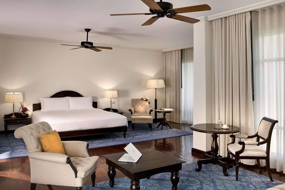 Ein Hotelzimmer mit einem Bett, zwei Stühlen, einem Couchtisch, einem Schreibtisch, einem Laptop und zwei Deckenventilatoren.