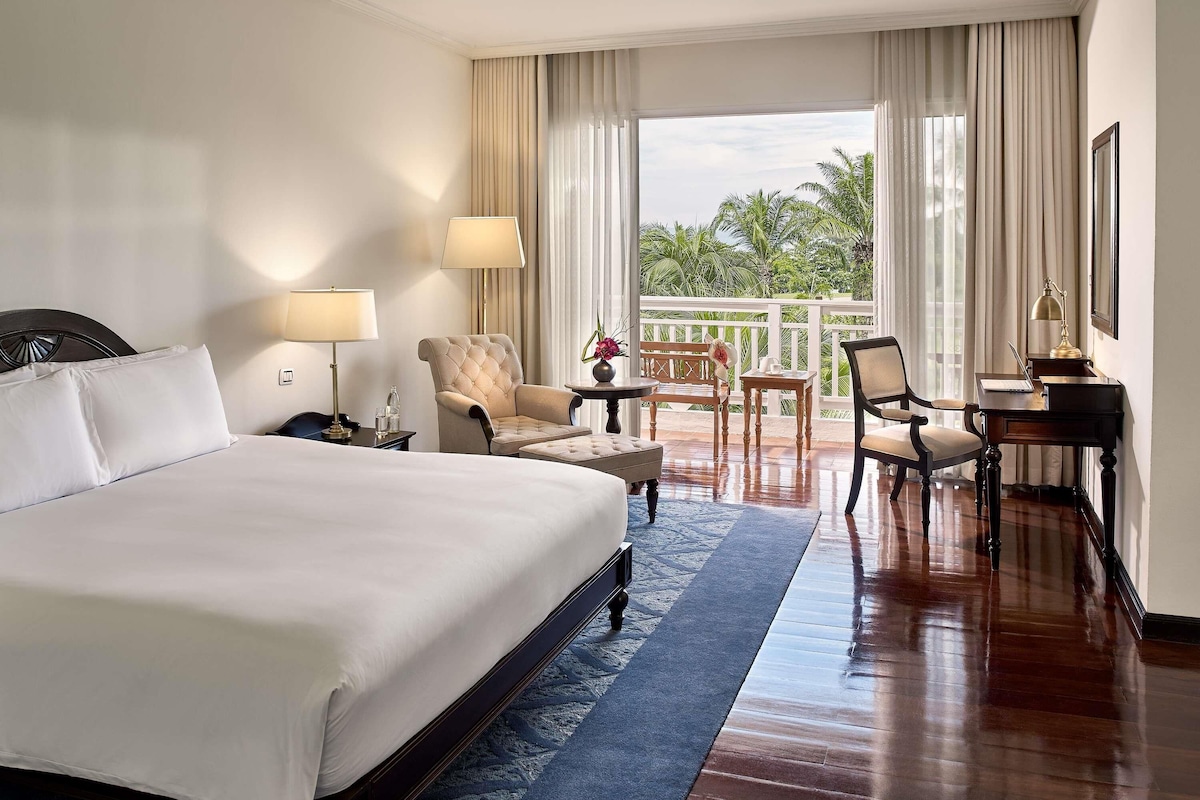 Habitación de hotel con una cama grande, dos sillas, un escritorio y un balcón con vistas a palmeras.