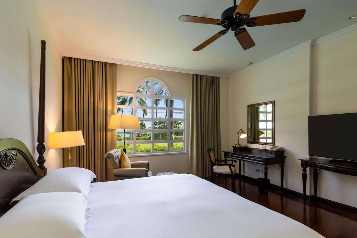 Ein Hotelzimmer mit einem großen Bett, einem Schreibtisch, einem Stuhl, einem Fernseher, einem Deckenventilator und einem Fenster mit Vorhängen.