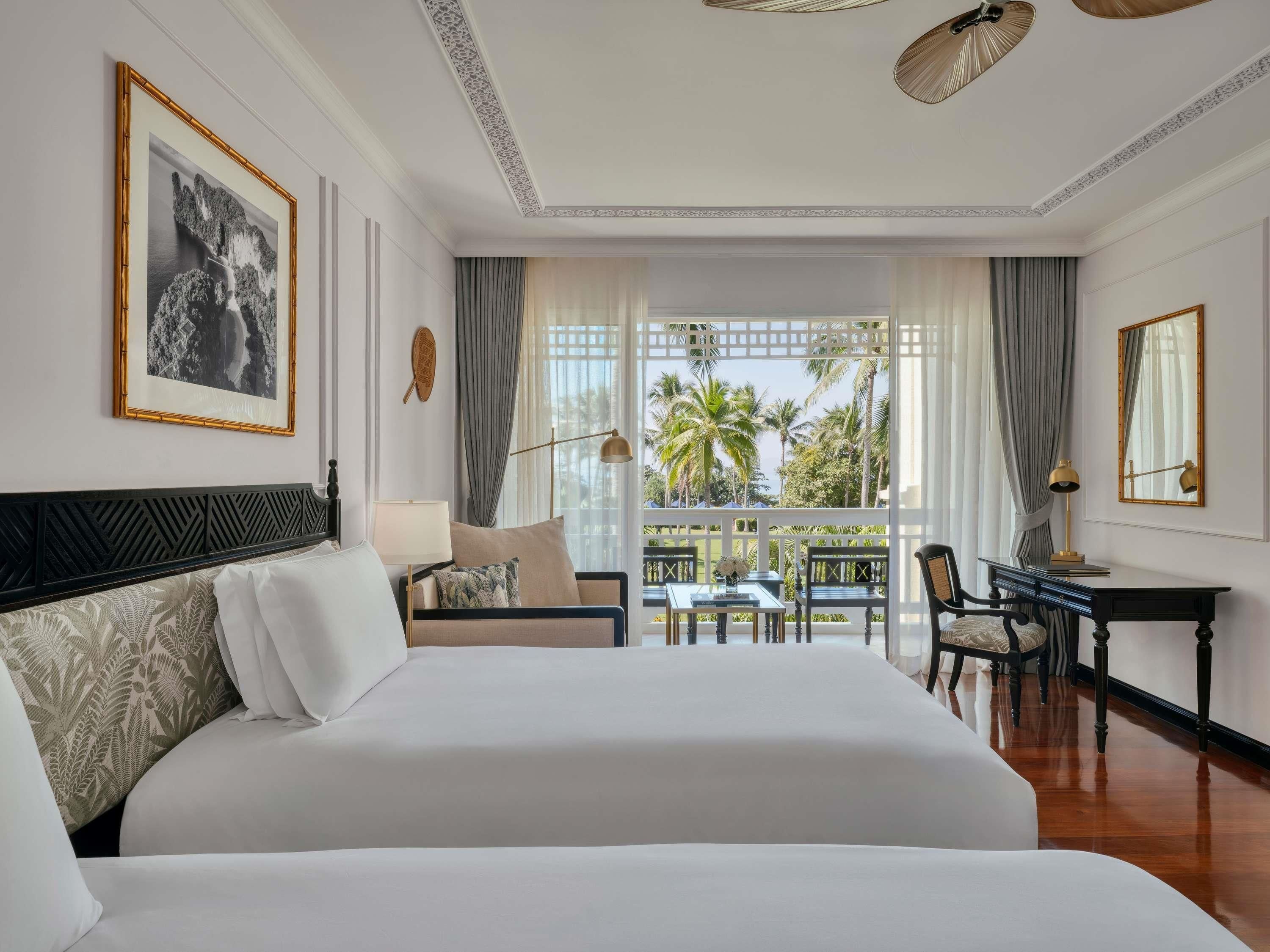 A hotel room with a large bed, a desk, a chair, a lamp, and a view of palm trees through the window.