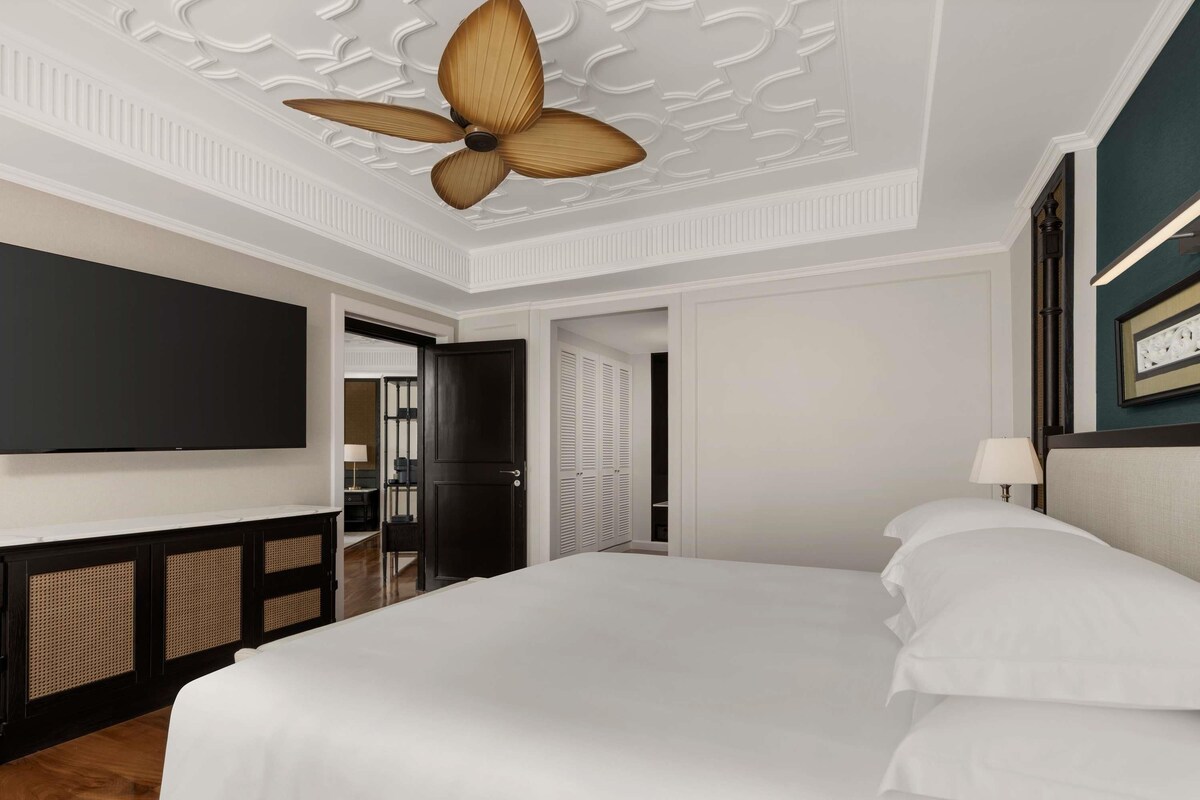 Une chambre d’hôtel avec un grand lit, un ventilateur de plafond, un téléviseur à écran plat et une porte donnant sur une salle de bain.