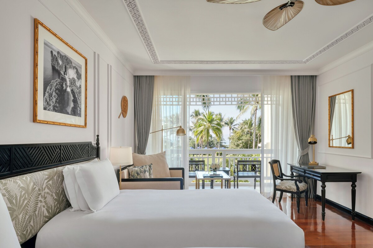 Habitación de hotel con una cama grande, un escritorio, una silla, una mesa y un balcón con vistas a palmeras.