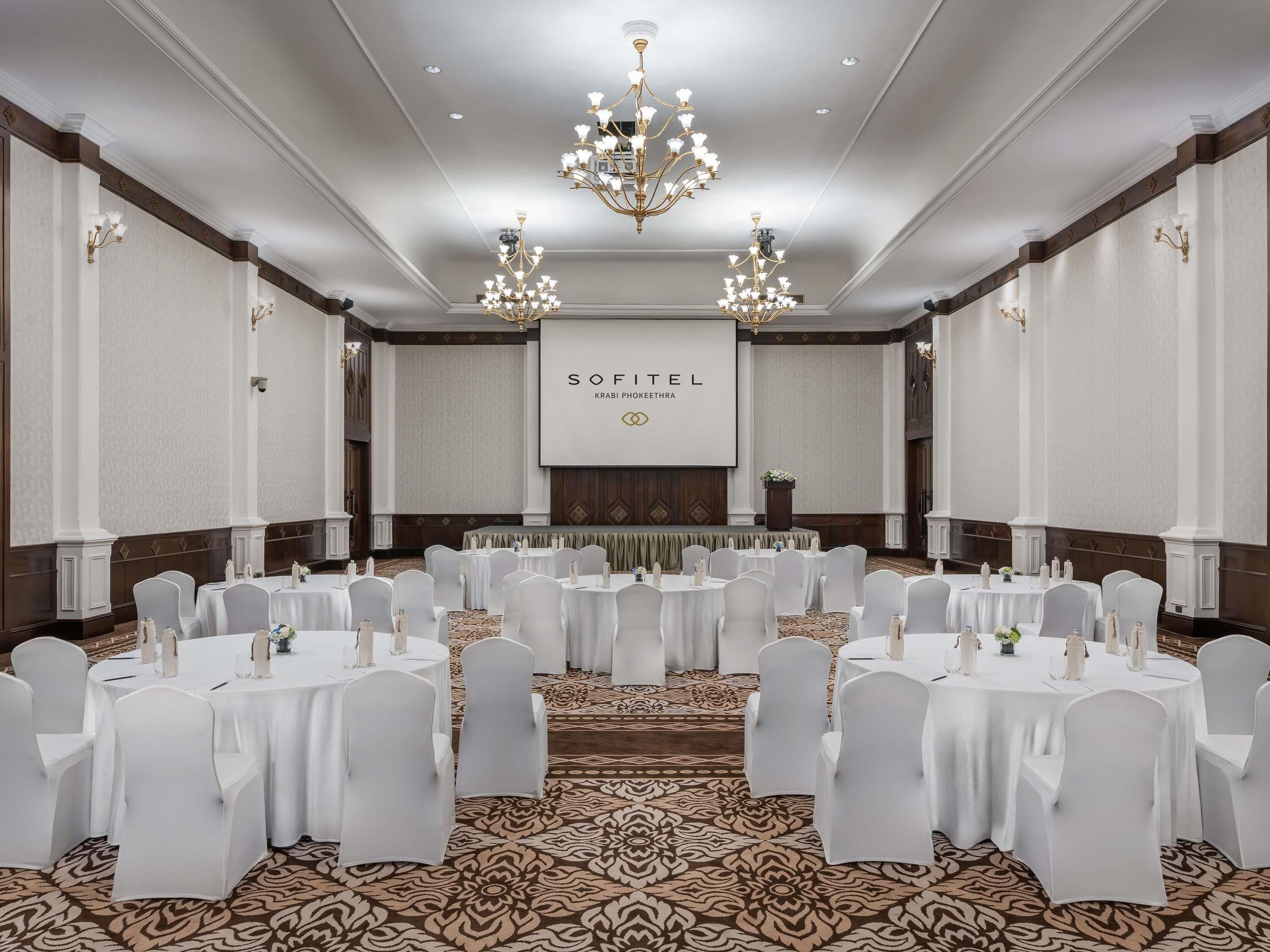 Un salón de conferencias amplio con mesas redondas, sillas blancas, candelabros y una pantalla de proyección que muestra 'SOFITEL'.