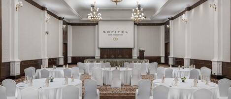 Un salón de conferencias amplio con mesas redondas, sillas blancas, candelabros y una pantalla de proyección que muestra 'SOFITEL'.