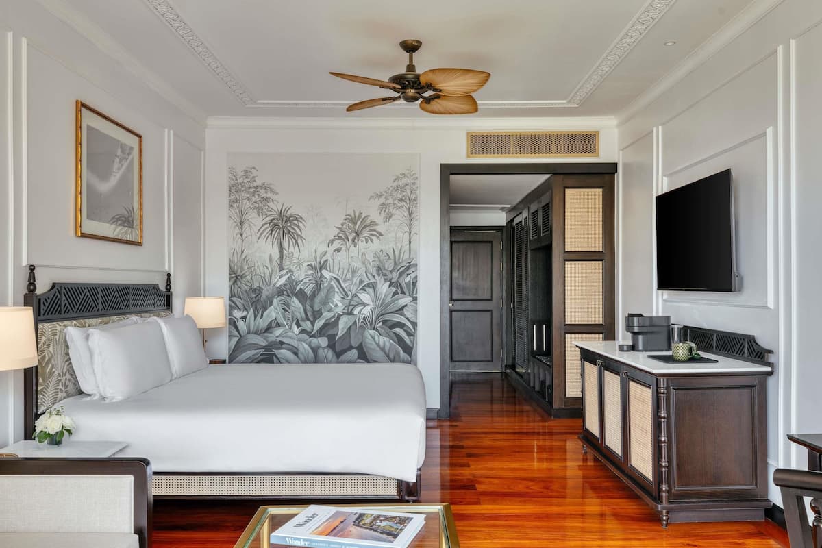 Une chambre dâhĂŽtel avec un lit, des lampes de chevet, un ventilateur de plafond, une grande fresque murale, un tĂ©lĂ©viseur, un minibar et un plancher en bois.