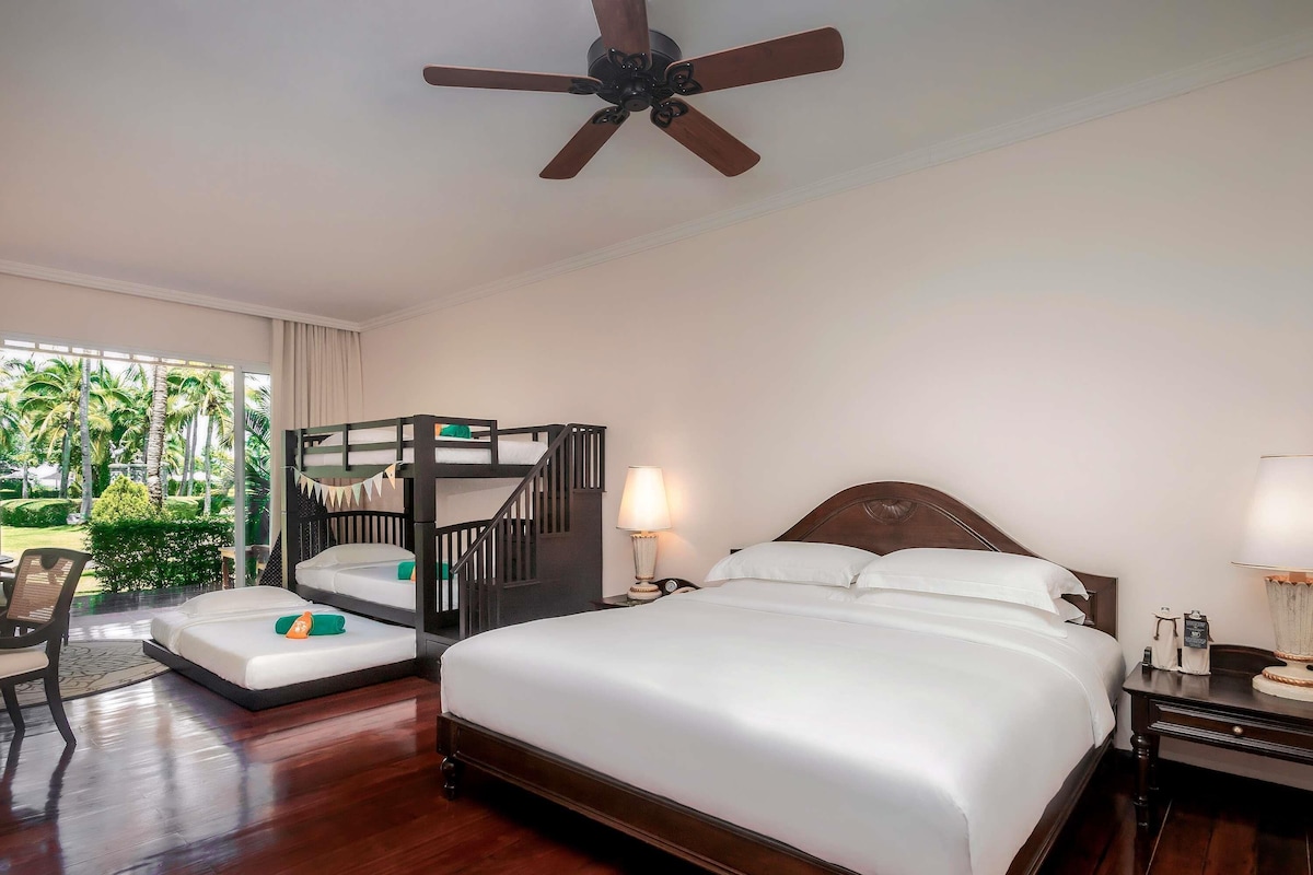 Habitación de hotel con una cama grande, una cama litera, un ventilador de techo, un escritorio con una silla y vistas a palmeras.