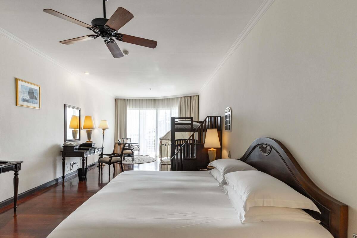 Habitación de hotel con una cama grande, un escritorio con lámparas, una silla, ventilador de techo y vistas al exterior a través del ventanal.