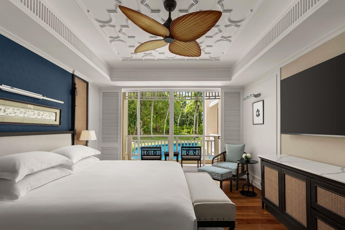 A hotel room with a large bed, a ceiling fan, a TV, and a view of a pool and greenery.