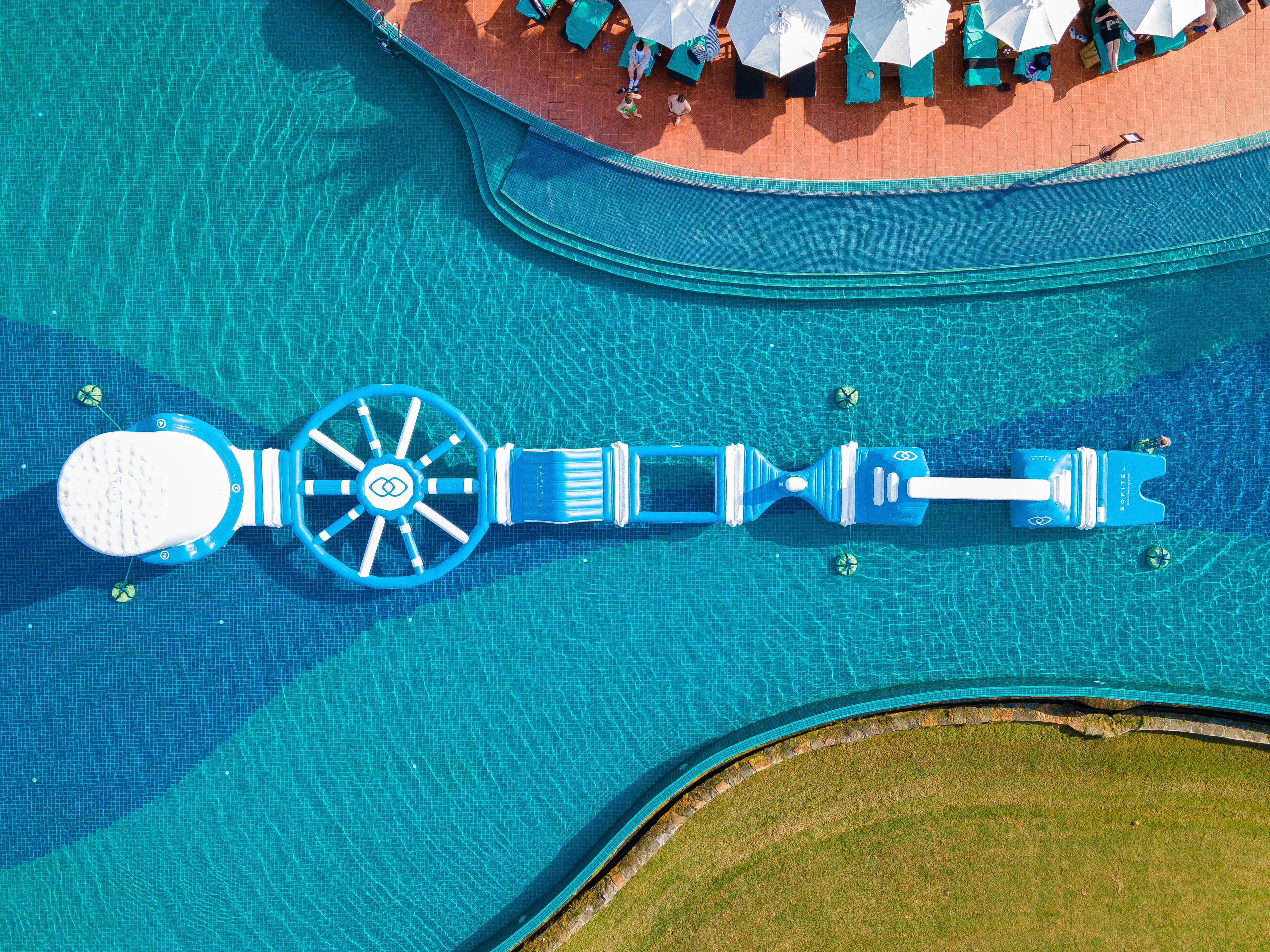 An aerial view of a swimming pool with a blue water slide, a lounge area with umbrellas, and a grassy area.