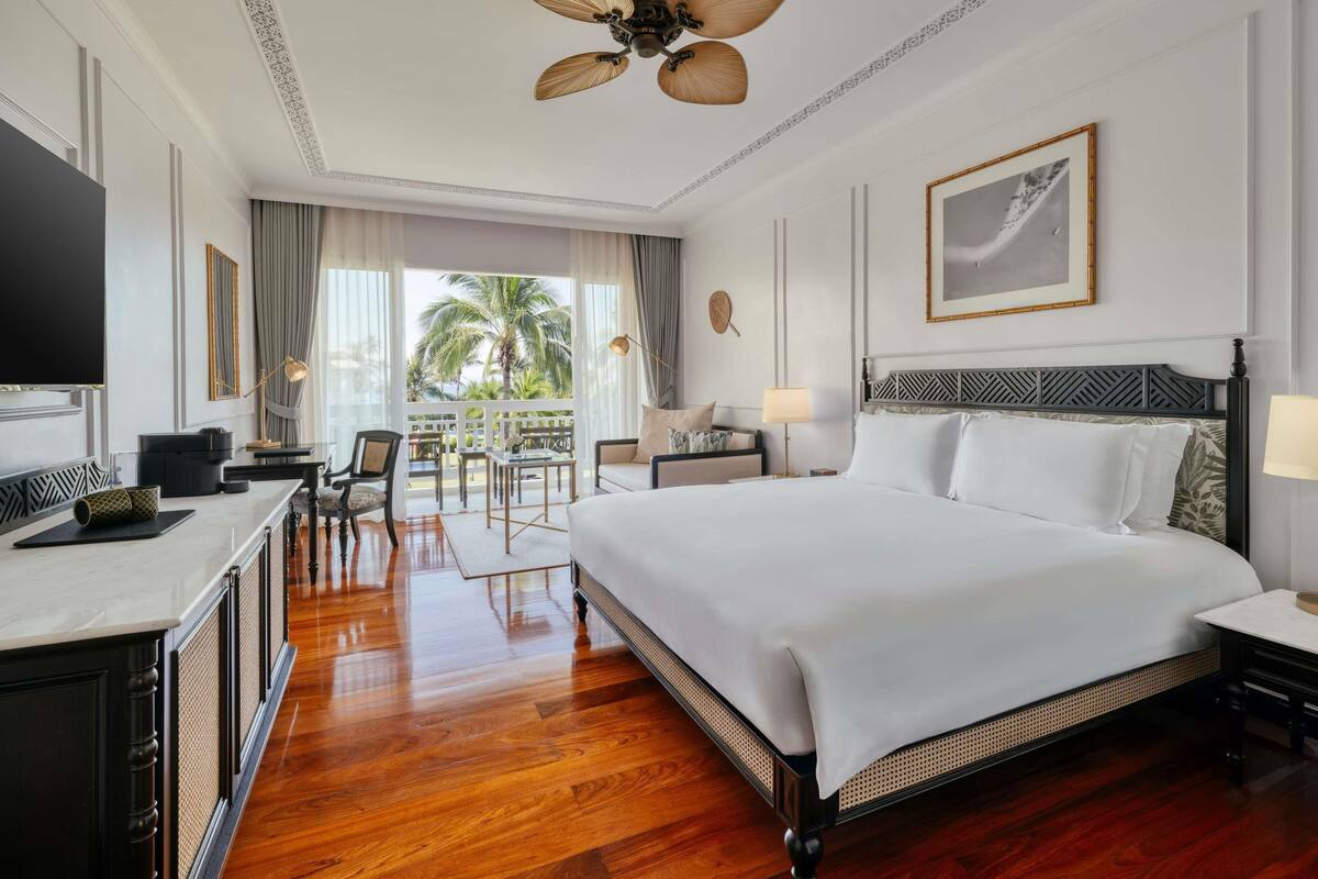 Ein Hotelzimmer mit einem großen Bett, einem Schreibtisch mit Flachbildfernseher, einem Balkon mit Blick auf Palmen und einem Deckenventilator.