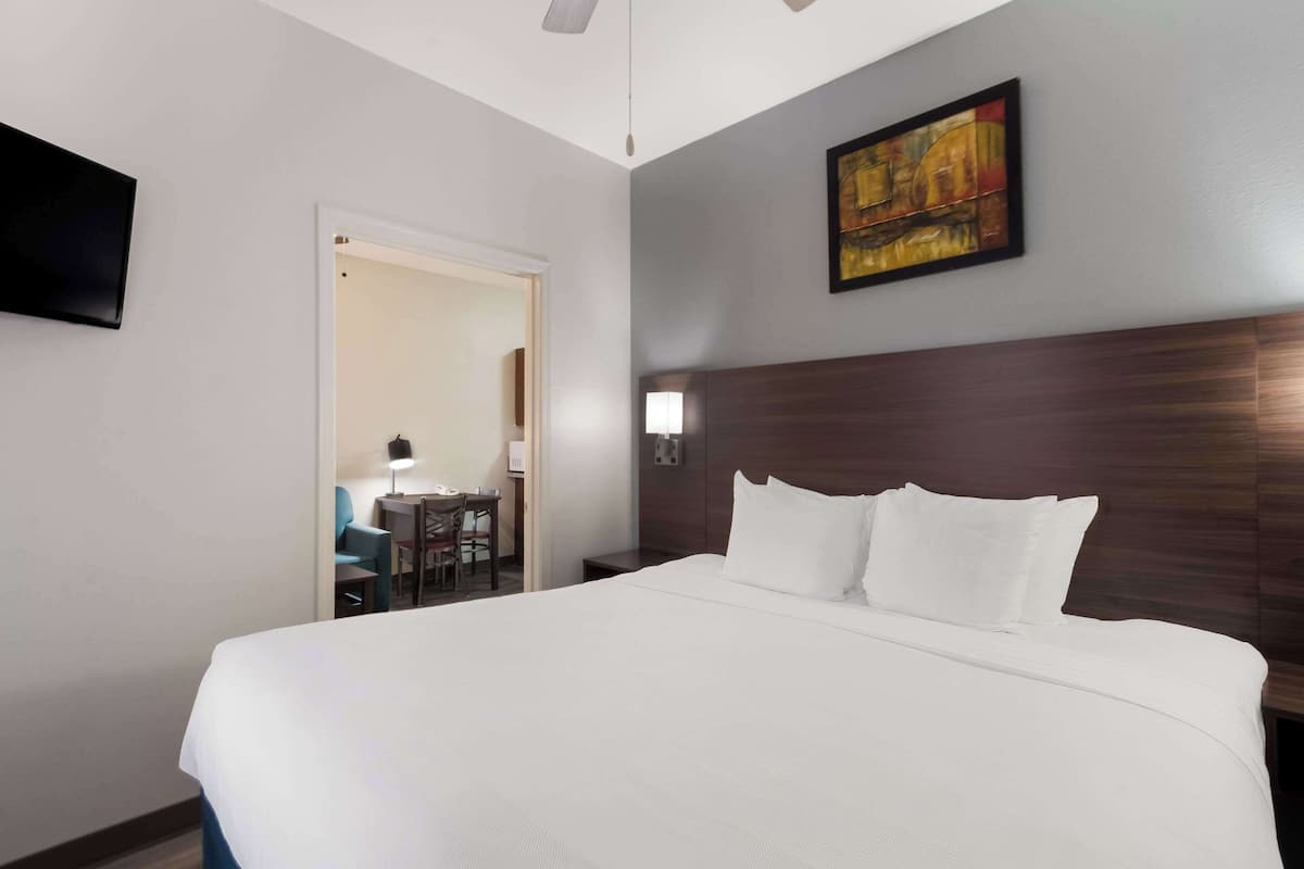 A hotel room with a large bed, a flat-screen TV mounted on the wall, a painting above the bed, and a desk with a chair and lamp in the corner.