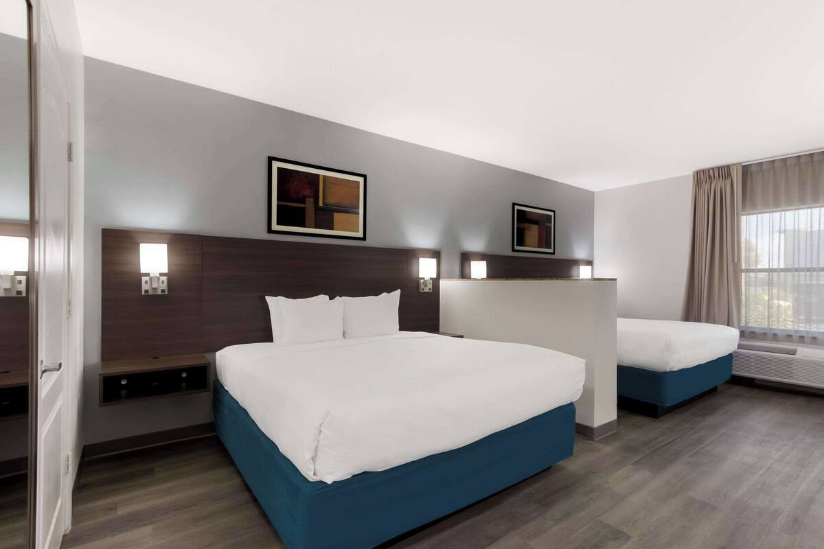 A hotel room with a large bed, a smaller bed, a nightstand, a wall-mounted TV, and a window with curtains.