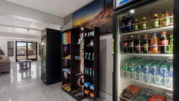 A vending machine stocked with various beverages and snacks, a refrigerator with bottled drinks, and a seating area with a view of the street.