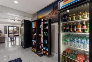 A vending machine stocked with various beverages and snacks, a refrigerator with bottled drinks, and a seating area with a view of the street. - MainStay Suites Extended Stay Hotel Casa Grande (Casa Grande)