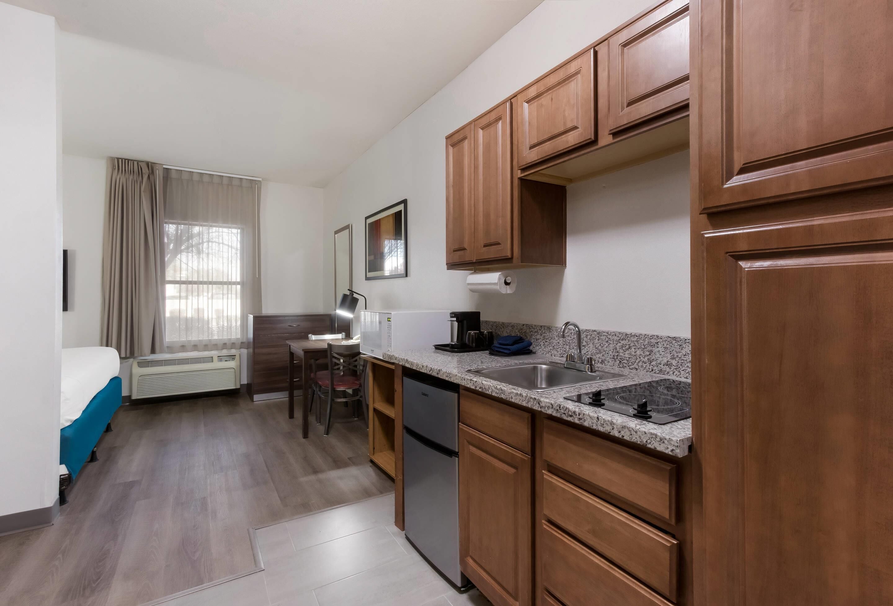 A compact hotel room with a kitchenette, a bed, a desk, and a window with curtains.