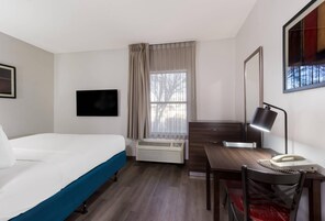 Habitación de hotel con cama, escritorio, silla, televisor, ventana con cortinas y radiador.