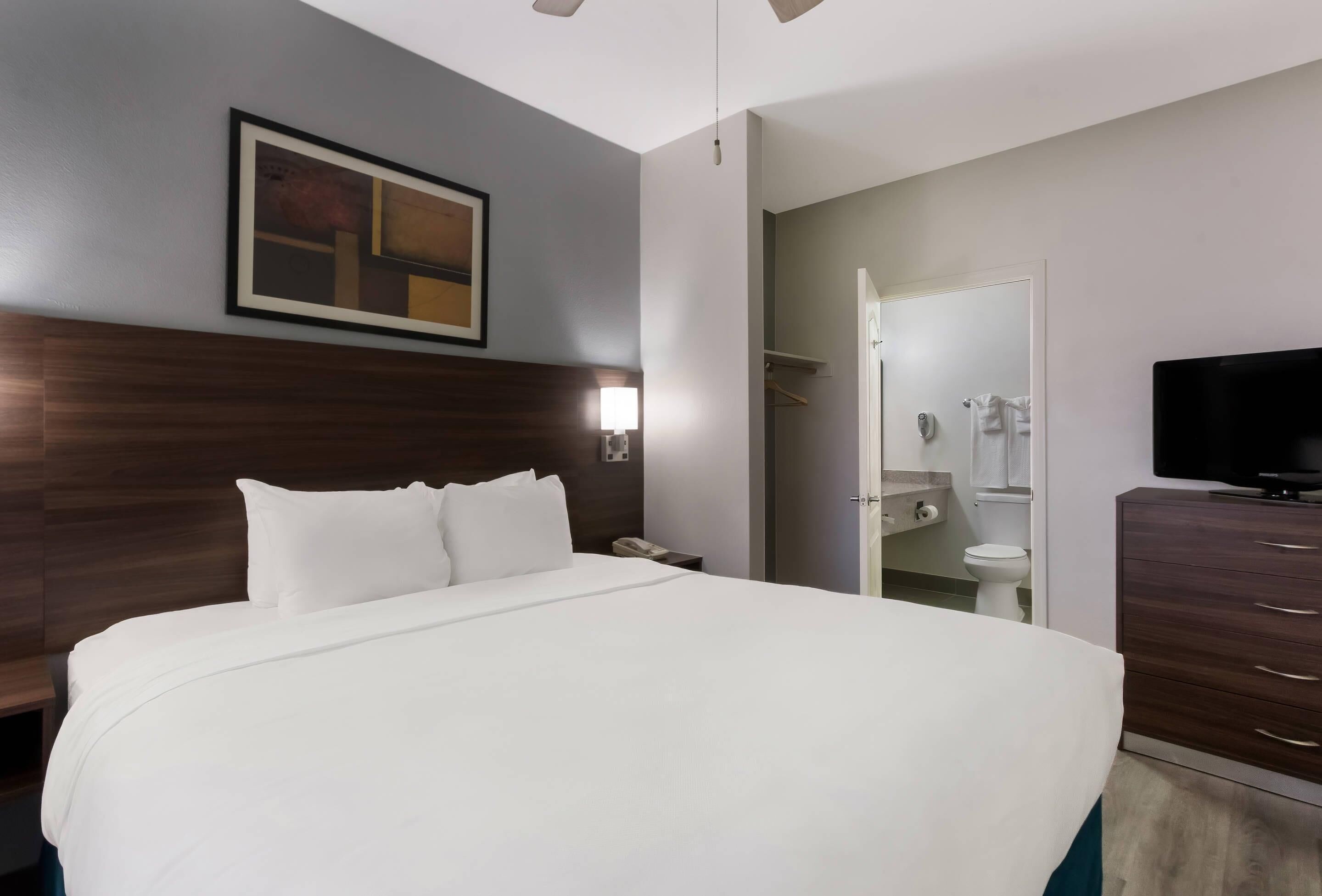A hotel room with a large bed, a bedside table, a lamp, a wardrobe, a television, and a bathroom visible through an open door.
