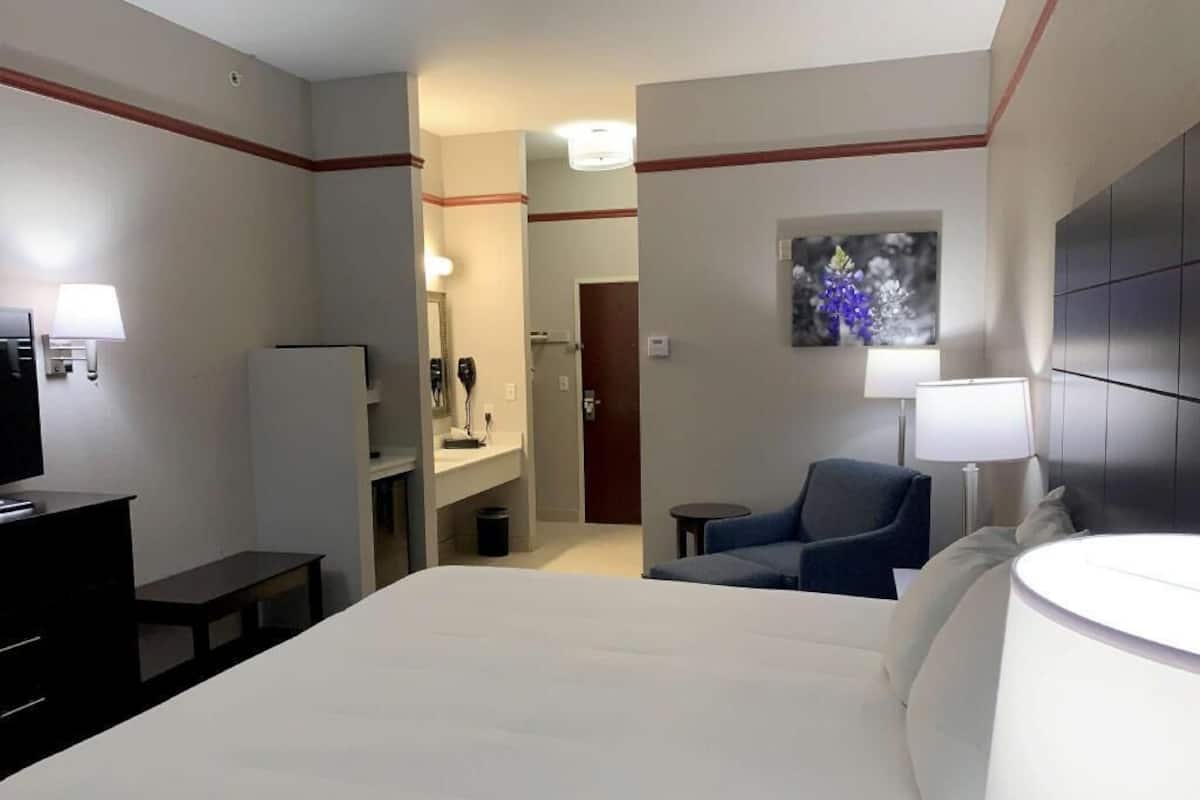A hotel room with a bed, a desk, a chair, a TV, a lamp, a painting, and a bathroom visible through an open door.