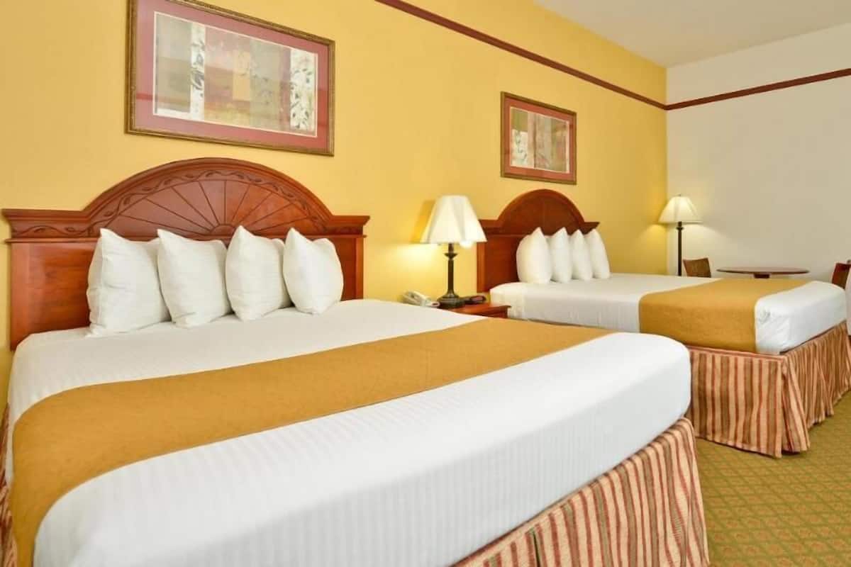 A hotel room with two beds, each with a wooden headboard, white bedding, and a striped skirt. There are two bedside tables with lamps, and the walls are painted yellow.