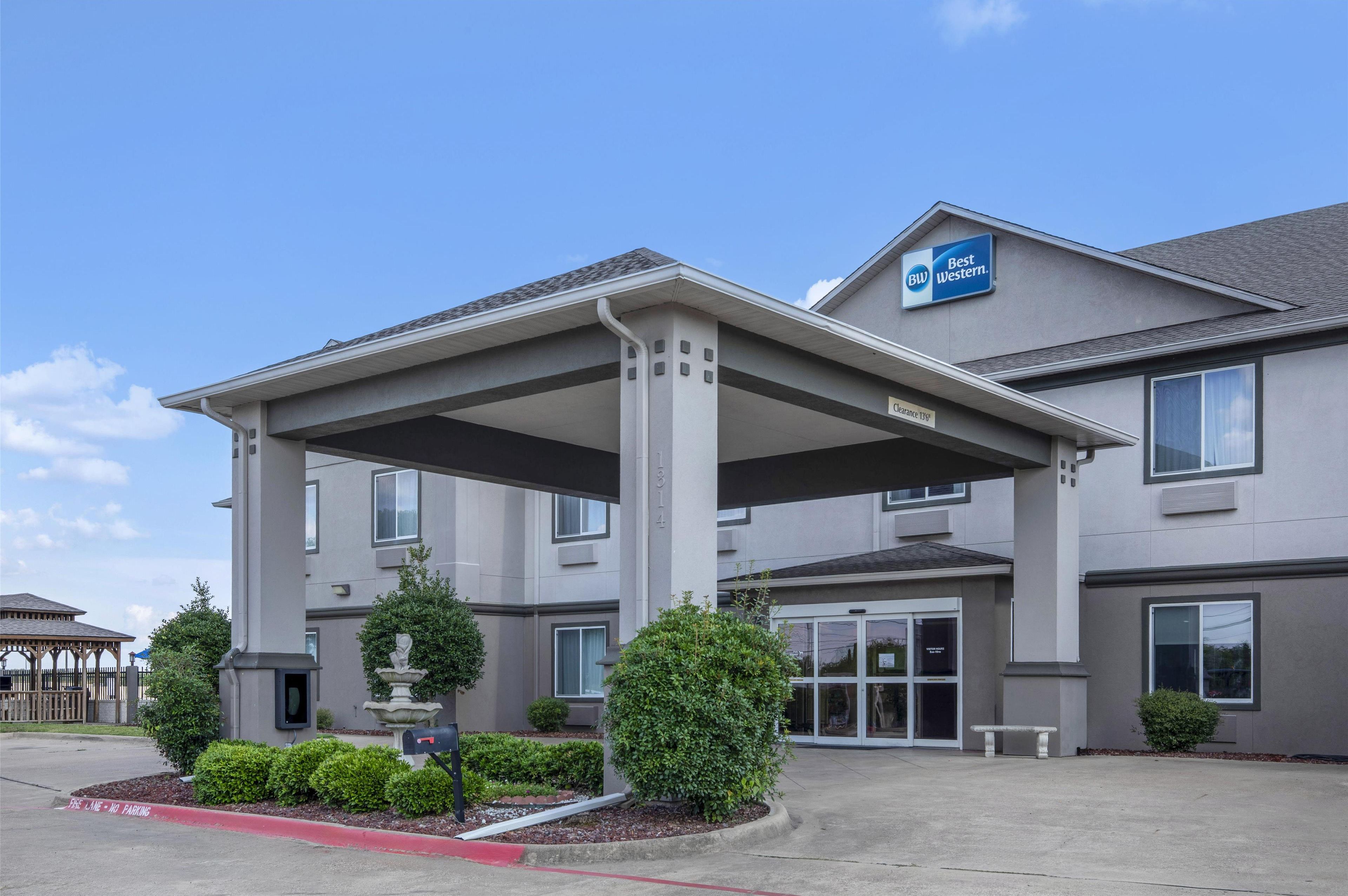 A Best Western hotel with a covered entrance, multiple windows, and a landscaped front yard with a fountain and trimmed bushes.