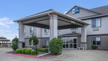 A Best Western hotel with a covered entrance, multiple windows, and a landscaped front yard with a fountain and trimmed bushes.