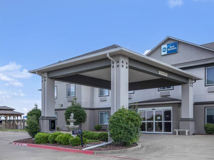 A Best Western hotel with a covered entrance, multiple windows, and a landscaped front yard with a fountain and trimmed bushes.
