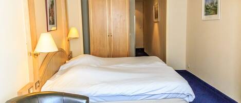 A hotel room with a bed, a chair, a desk, a lamp, and a wardrobe.