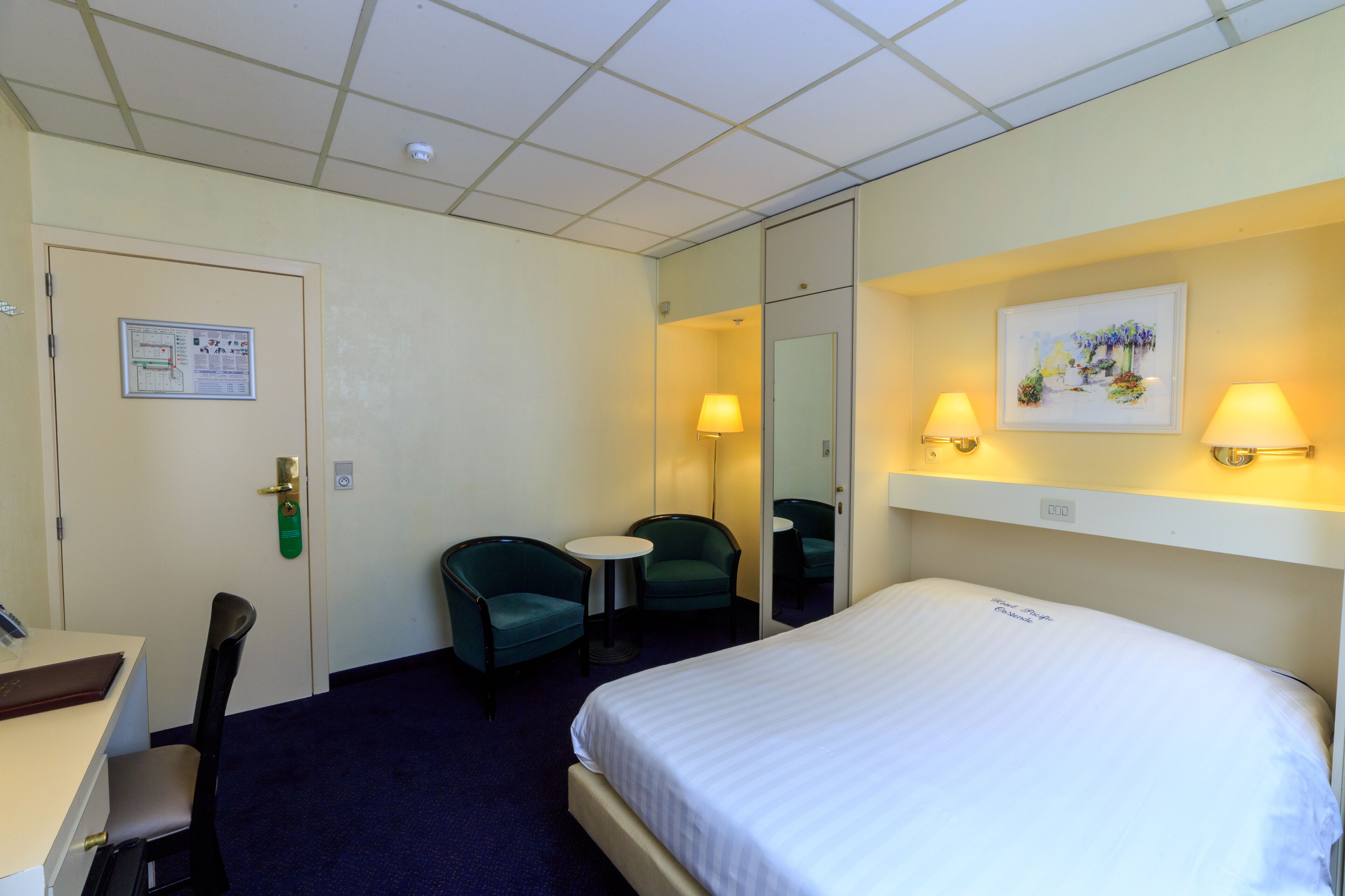 Et hotelværelse med en seng, to stole, et skrivebord, et lille bord, et spejl og et billede på væggen.
