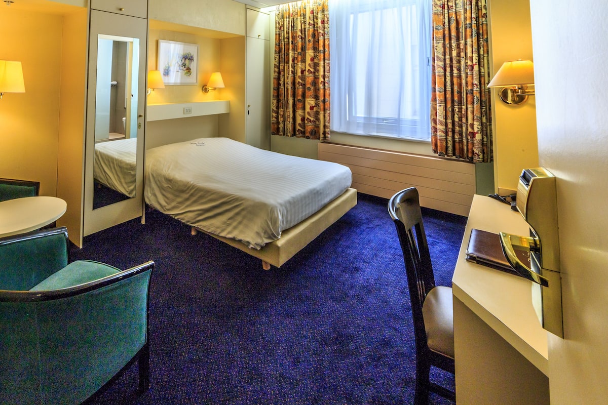 A hotel room with a bed, a desk, a chair, a mirror, and a window with curtains.