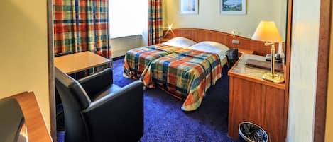 Una habitación de hotel con cama, escritorio, silla, lámpara y cubo de basura.