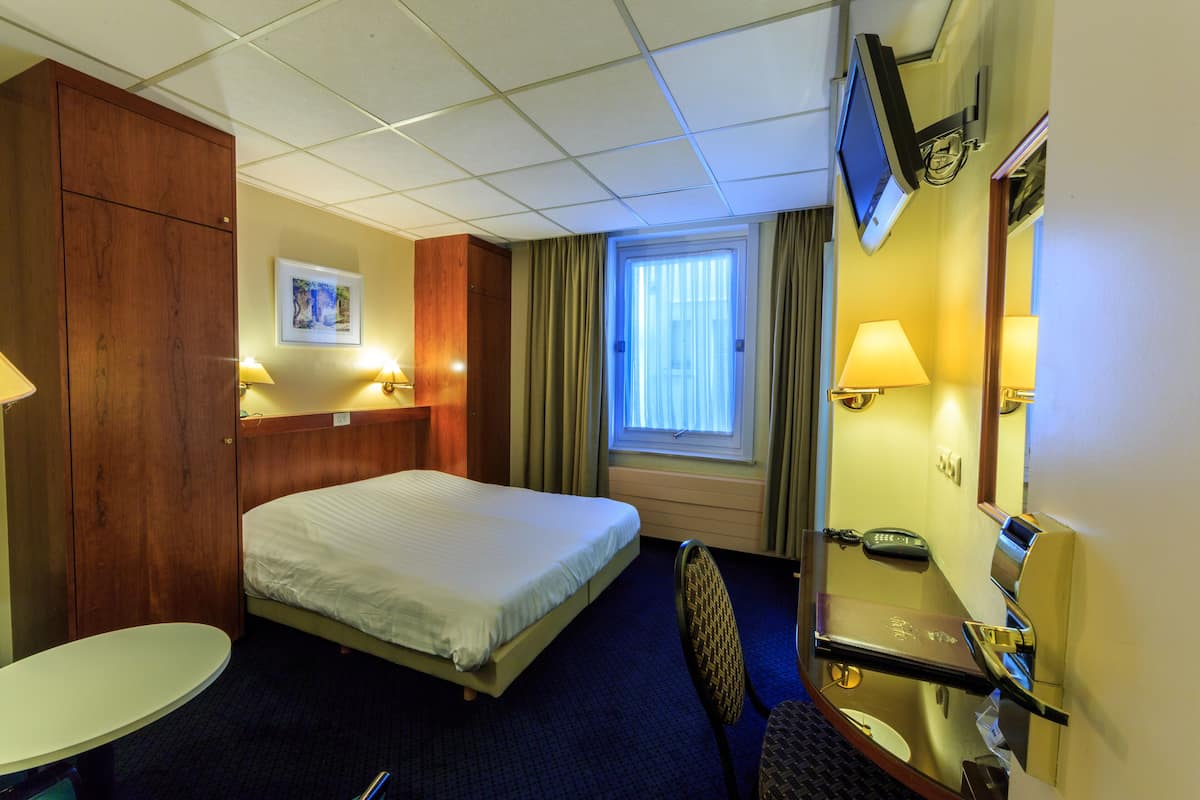 A hotel room with a bed, a desk with a chair, a TV mounted on the wall, and a window with curtains.