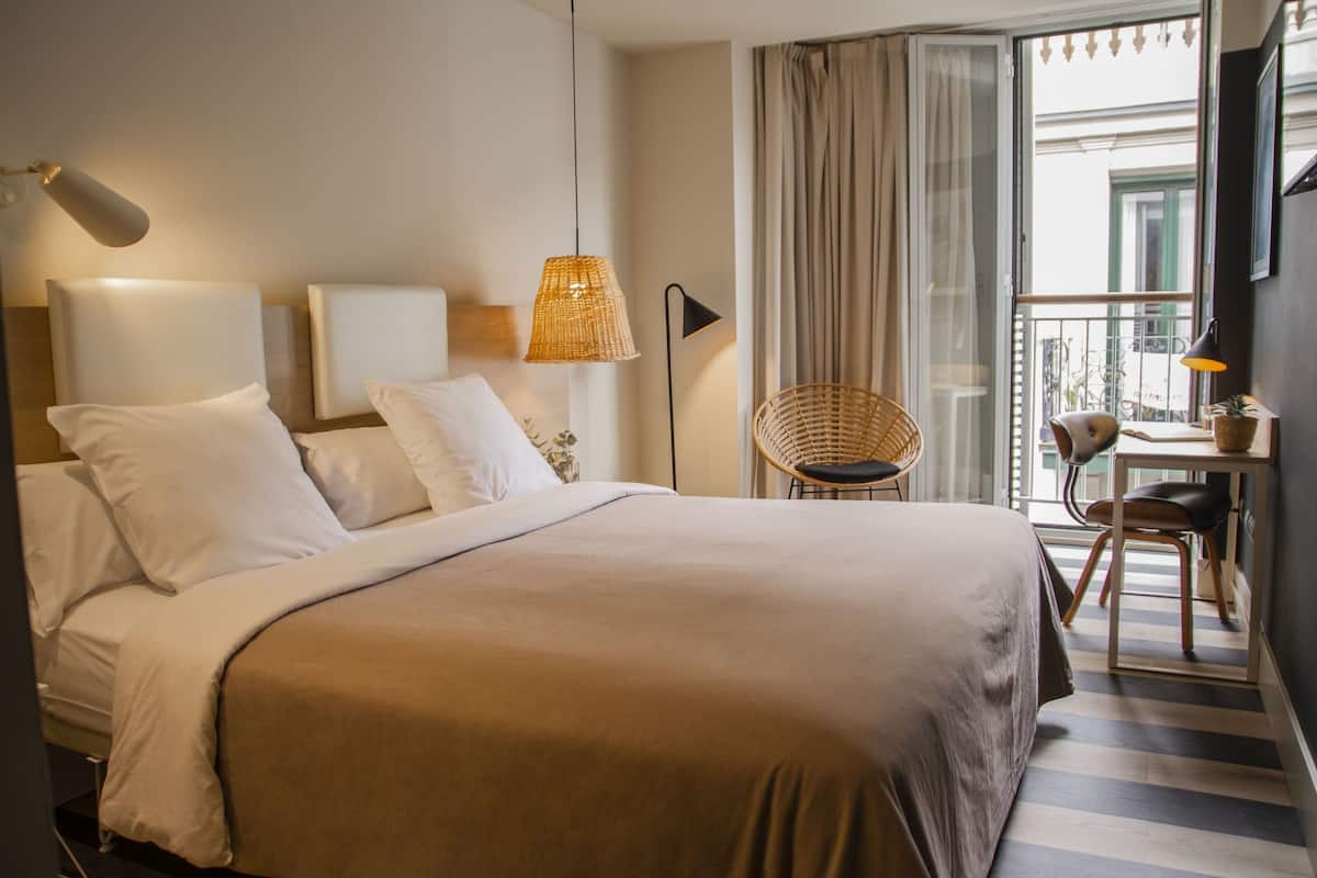 A hotel room with a large bed, a desk with a chair, a wicker chair, and a view of the outside through a window.