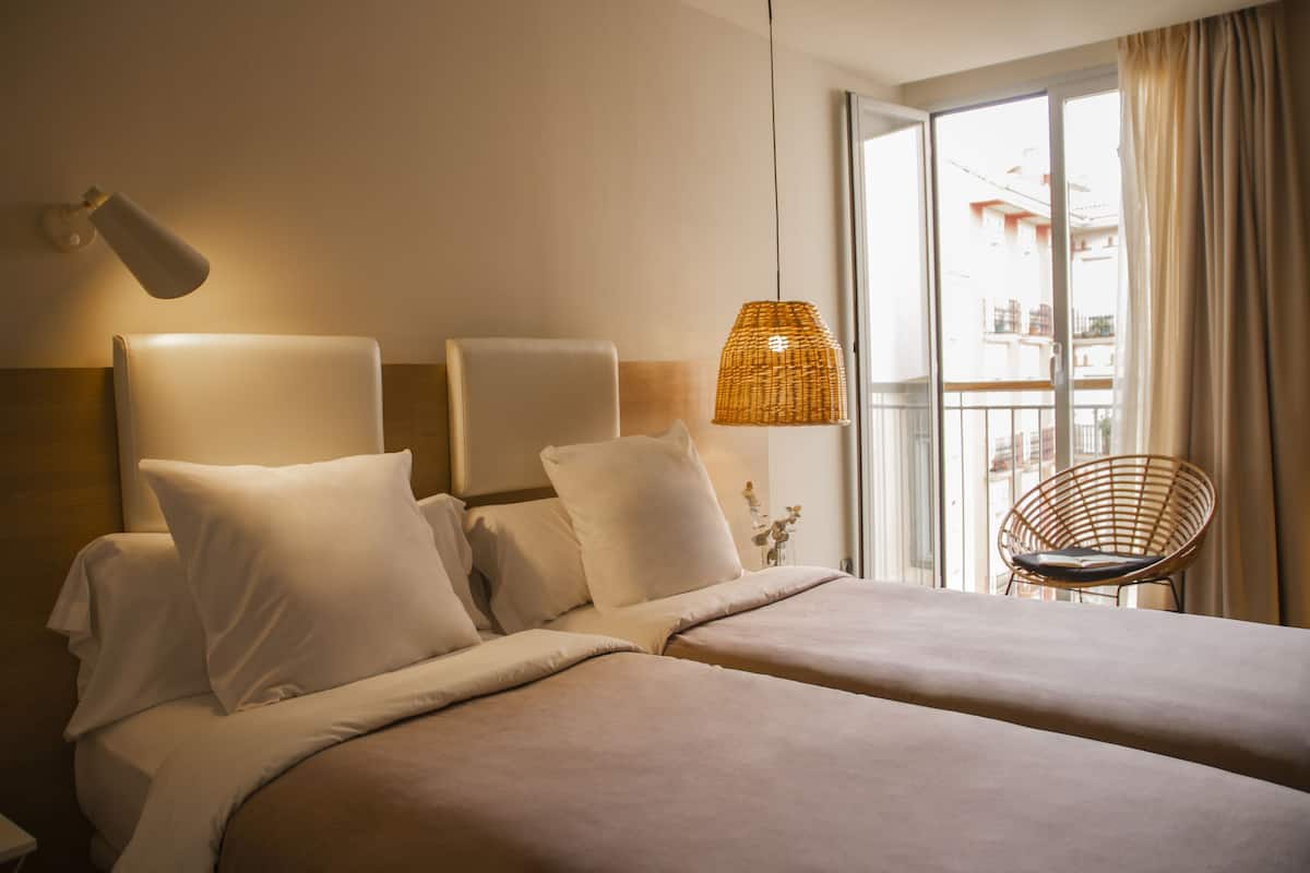 A hotel room with two beds, a wicker lamp, and a chair by the window.